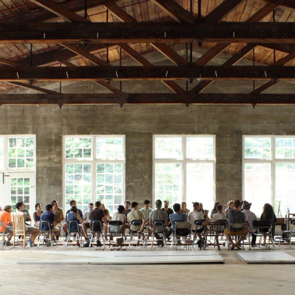 large lofted cieilng building with open floor plan and large windows. About 40 people are seated in a circle with a speaker at the front of the room