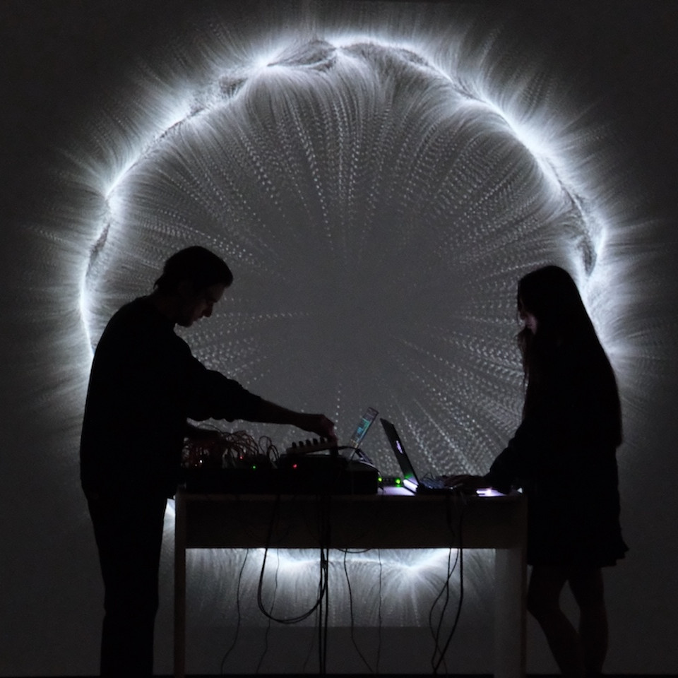 Two audio/visual performers standing in front of their projected artwork. BLCK SUN / Amiangelika/ Multimedia Performance / CURRENTS 2024 @ El Museo Cultural