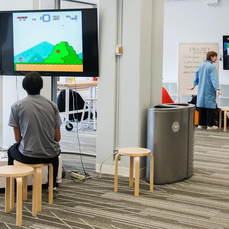 Students in a brightly lit collaborative working space. A student with his back to the camera is playing Super Mario Brothers.