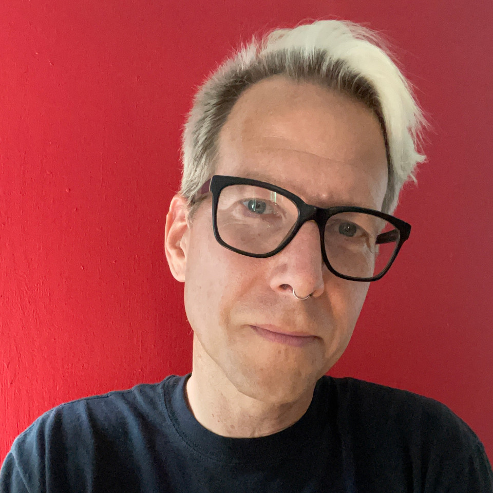 Headshot of Rob Ray. Bleached white short hair and black glasses, with black t-shirt